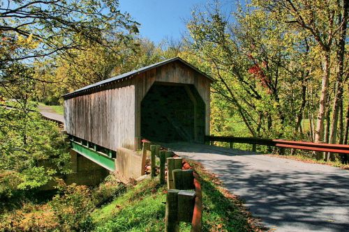A wooden covered bridge spans a small road, surrounded by trees with autumn foliage under a clear blue sky.