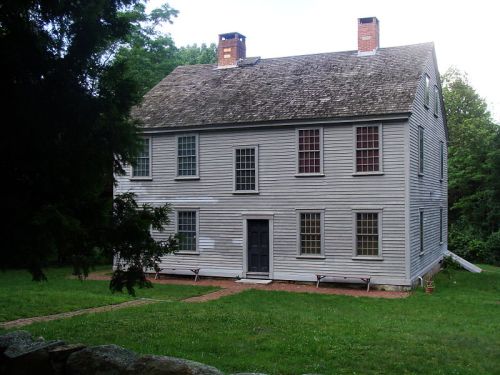 A two-story gray wooden house with a steep roof, surrounded by grass and trees, featuring multiple windows and a front door.