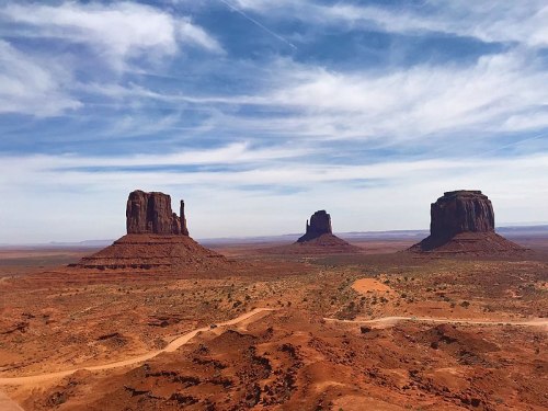 A scenic view of Monument Valley featuring three iconic rock formations against a blue sky with wispy clouds.