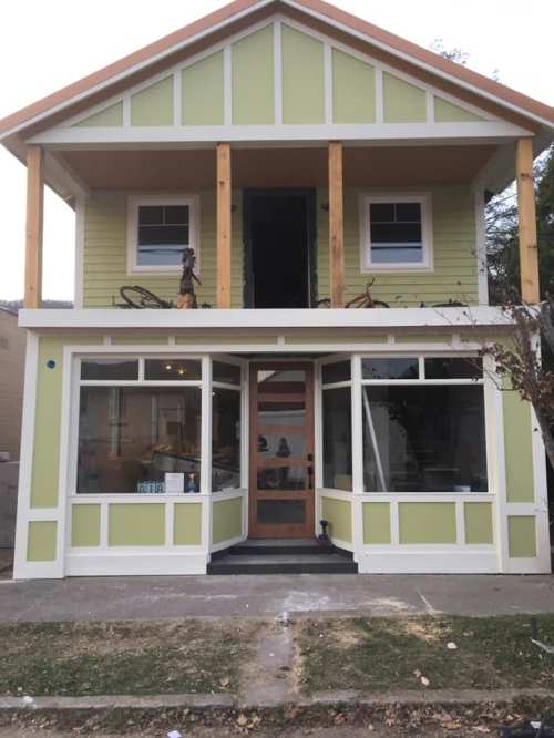 A two-story house with green siding, large windows, and wooden accents, under construction with an open front.