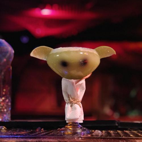A green cocktail with a rimmed glass, resembling a character with ears, set against a colorful, dimly lit background.