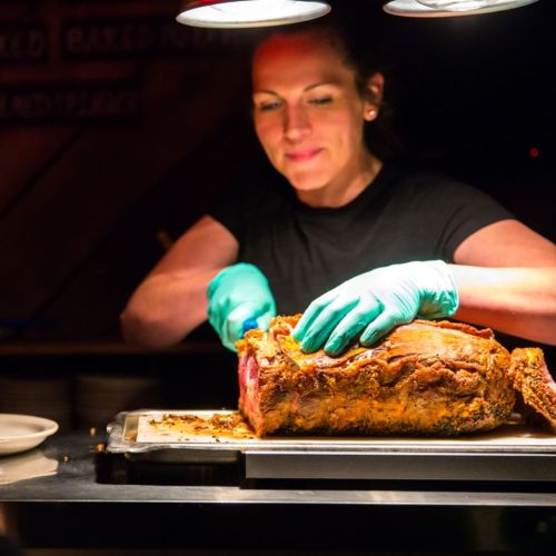 A chef in gloves carves a large roast on a cutting board under warm lighting.