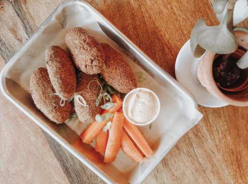 A tray with crispy croquettes, carrot sticks, and a small cup of dipping sauce, alongside a potted plant.