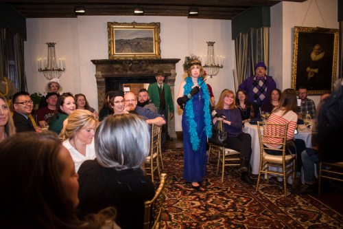 A lively dinner scene with guests seated at tables, while a performer in a blue dress and feather boa entertains.