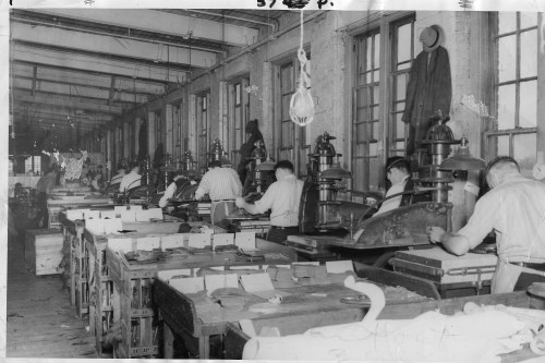 Black and white photo of workers operating machinery in a large industrial workshop with tables and windows.
