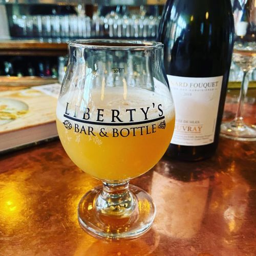 A glass of beer sits on a copper bar top, with a bottle of wine in the background at Liberty's Bar & Bottle.