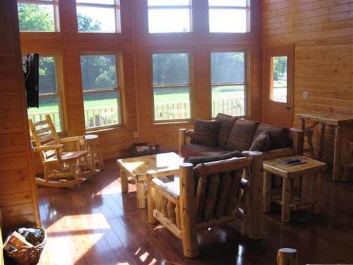 Cozy wooden living room with large windows, log furniture, and a view of greenery outside.