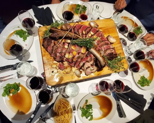 A large platter of sliced steak garnished with herbs, surrounded by plates, glasses of wine, and fries on a dining table.