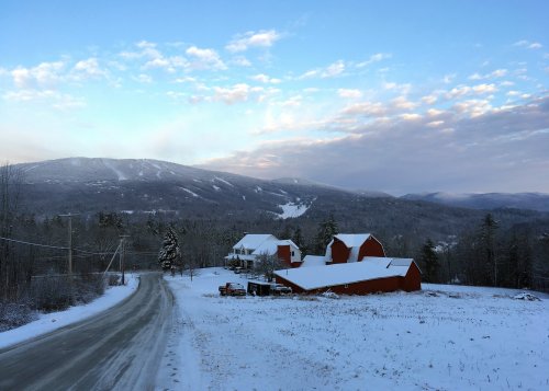 A snowy landscape featuring red barns, a winding road, and mountains under a blue sky with scattered clouds.