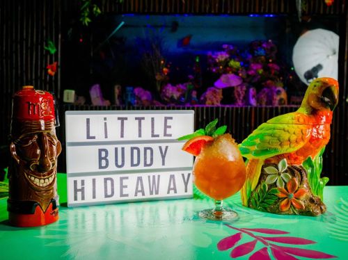 A colorful cocktail sits on a table next to a tiki mug and a parrot statue, with a sign reading "Little Buddy Hideaway."