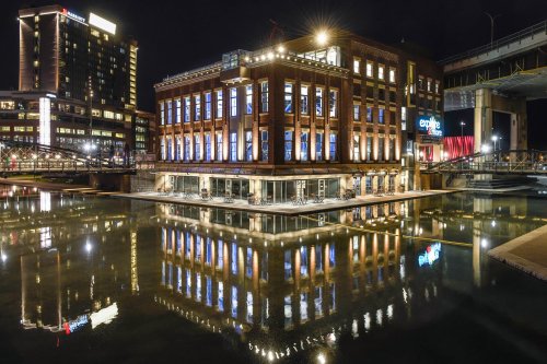 A historic building at night, reflecting in calm water, surrounded by modern city lights and structures.