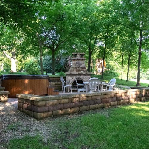 A cozy outdoor space featuring a hot tub, stone fireplace, and seating area surrounded by lush greenery.