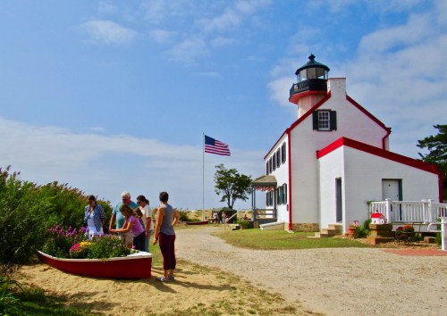 Visitors admire flowers near a lighthouse, with an American flag waving in the background on a sunny day.