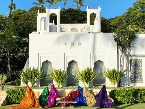 A vibrant scene with people in colorful, shimmering outfits seated in front of a white building surrounded by greenery.