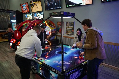 Four friends play air hockey at an arcade, with colorful games and screens in the background.