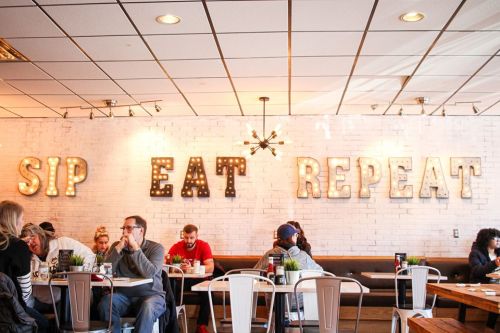 A cozy restaurant interior with a "Sip Eat Repeat" sign, featuring diners at tables and a bright, inviting atmosphere.