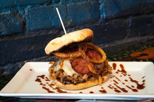 A hearty sandwich with pulled pork, sausage, cheese, and crispy onion rings, served on a plate with a decorative sauce.