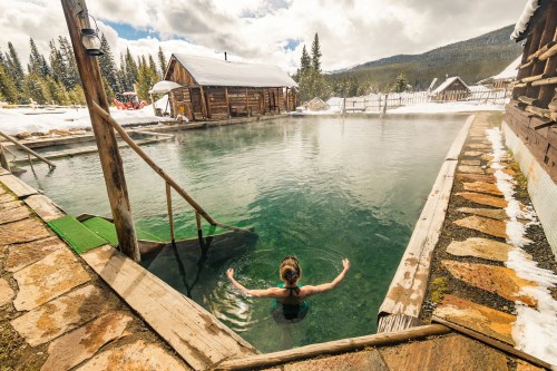 A person relaxes in a natural hot spring surrounded by snow-covered trees and rustic wooden cabins.