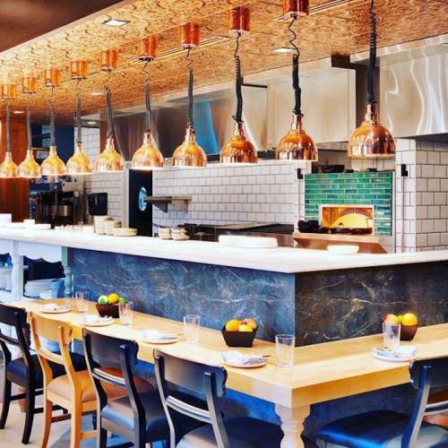 Modern restaurant interior featuring a sleek bar, copper pendant lights, and a visible pizza oven in the background.
