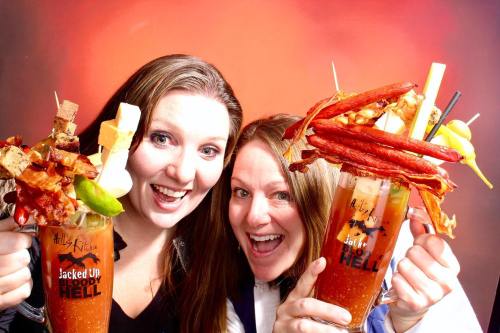 Two women joyfully hold oversized Bloody Mary cocktails topped with various garnishes, smiling at the camera.