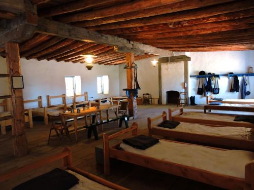 Interior of a rustic room with wooden beams, featuring simple beds, a dining table, and a fireplace.