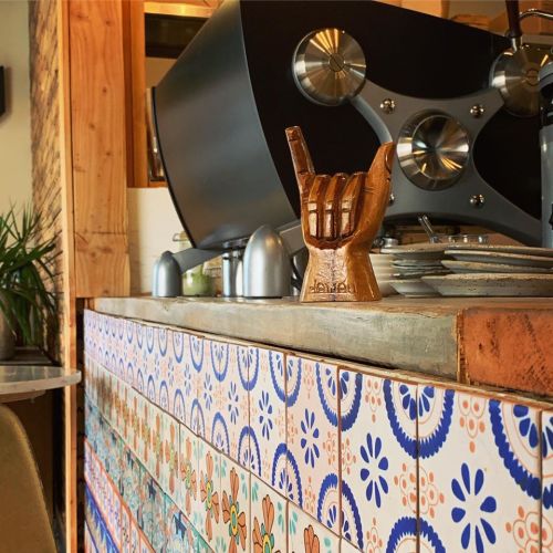 A wooden hand sculpture making a "shaka" gesture on a tiled counter in a cozy café setting.