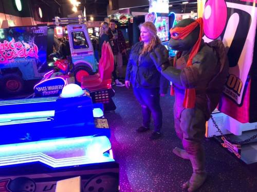 A woman stands next to a costumed character resembling a turtle in an arcade, with colorful game machines in the background.