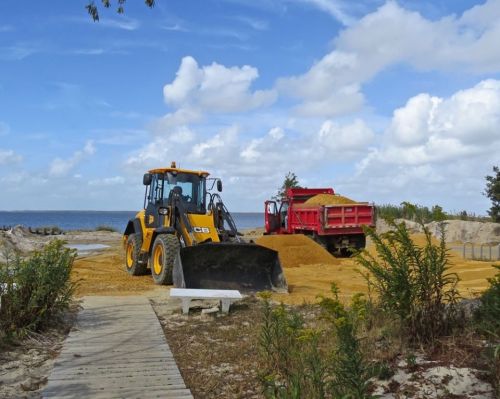 A yellow backhoe loader and a red dump truck are on a sandy beach under a blue sky with fluffy clouds.