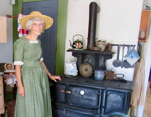 A woman in a green dress and straw hat stands beside a vintage black stove in a rustic kitchen setting.