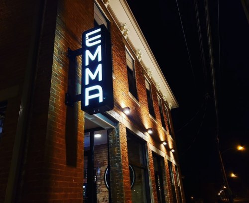 A brick building at night with a bright sign reading "EMMA" illuminated above the entrance.