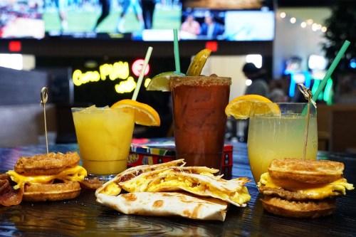A table with breakfast items: waffles with bacon and eggs, a burrito, and colorful cocktails in a lively setting.