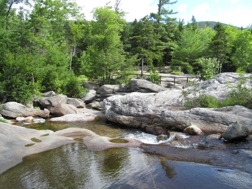 A serene landscape featuring a rocky stream surrounded by lush green trees and a clear blue sky.