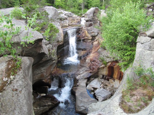 A serene waterfall cascades over rocks into a clear pool, surrounded by lush greenery and rugged stone formations.