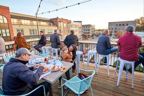 A rooftop dining area with people enjoying food and drinks, overlooking a lively street at sunset.