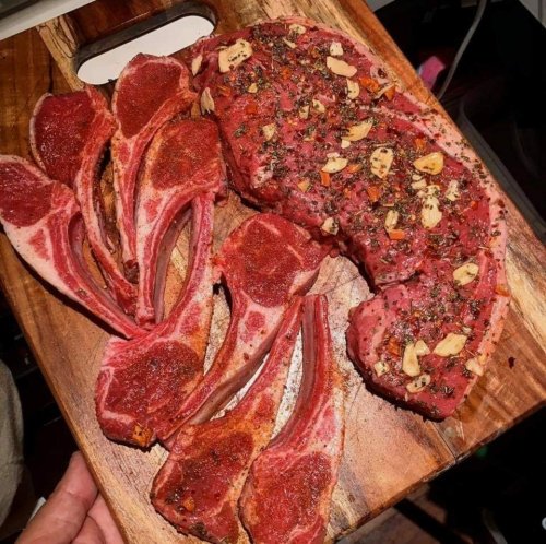 A wooden cutting board with seasoned lamb chops and a large rib chop, garnished with garlic and herbs.