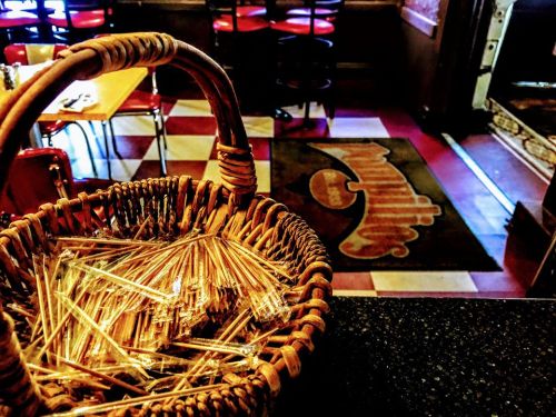 A woven basket filled with wooden toothpicks, with a restaurant interior and patterned rug in the background.