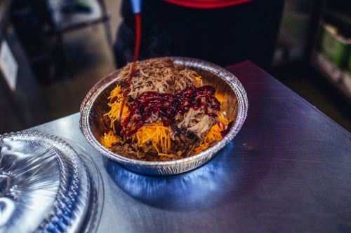 A metal bowl filled with shredded meat, shredded cheese, and drizzled with barbecue sauce.