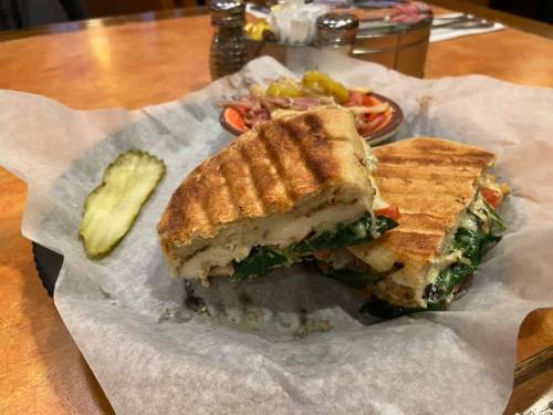 A grilled sandwich with chicken, spinach, and tomatoes, served with a pickle and a side of salad on a plate.