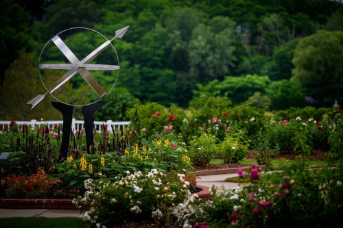 A modern metal sculpture with arrows stands in a vibrant garden filled with colorful flowers and lush greenery.