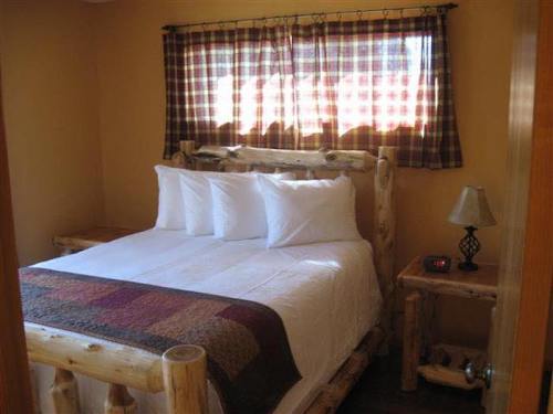 Cozy bedroom with a log bed, white bedding, plaid curtains, and a bedside lamp. Warm, inviting atmosphere.