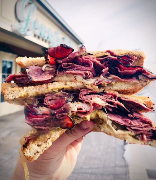 A hand holding a stacked sandwich filled with layers of pastrami and mustard, with a blurred restaurant sign in the background.