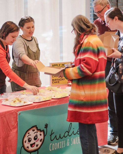 A colorful event scene with people sampling cookies at a table, featuring a banner for "Tudie's Cookies & Sweets."