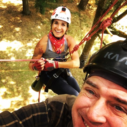 Two people smiling while climbing trees, wearing helmets and harnesses, with ropes and trees in the background.