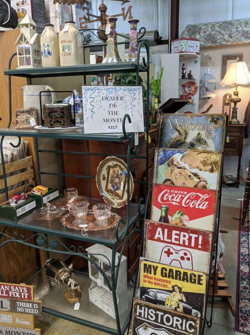 A display shelf with vintage items, including glassware and signs, labeled "Dealer of the Month" in a shop setting.