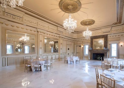 Elegant banquet hall with chandeliers, mirrors, and round tables set for dining, featuring marble floors and ornate decor.
