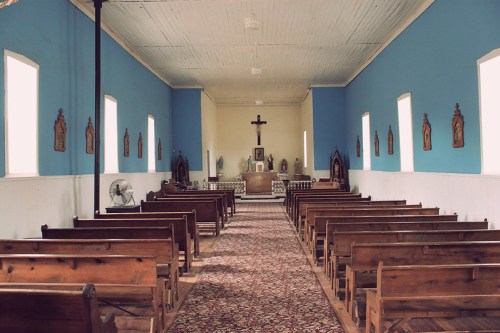 Interior of a simple church with wooden benches, blue walls, and a central altar featuring a cross and religious icons.