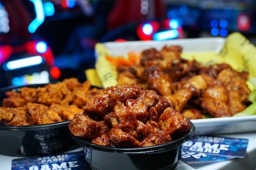 A close-up of two bowls of chicken wings, one spicy and one BBQ, with colorful arcade games in the background.