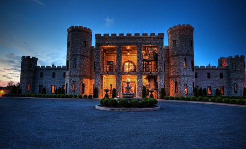A grand stone castle with turrets, illuminated at dusk, featuring a central fountain and manicured landscaping.