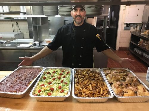 A chef stands behind four large trays of food, including salad, fries, and roasted chicken, in a kitchen setting.