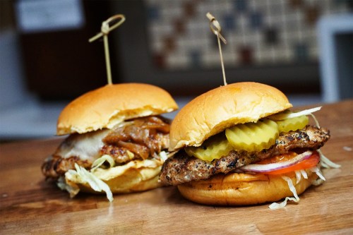 Two burgers on a wooden surface, one with grilled chicken, pickles, lettuce, and tomato, the other with pulled pork and onions.